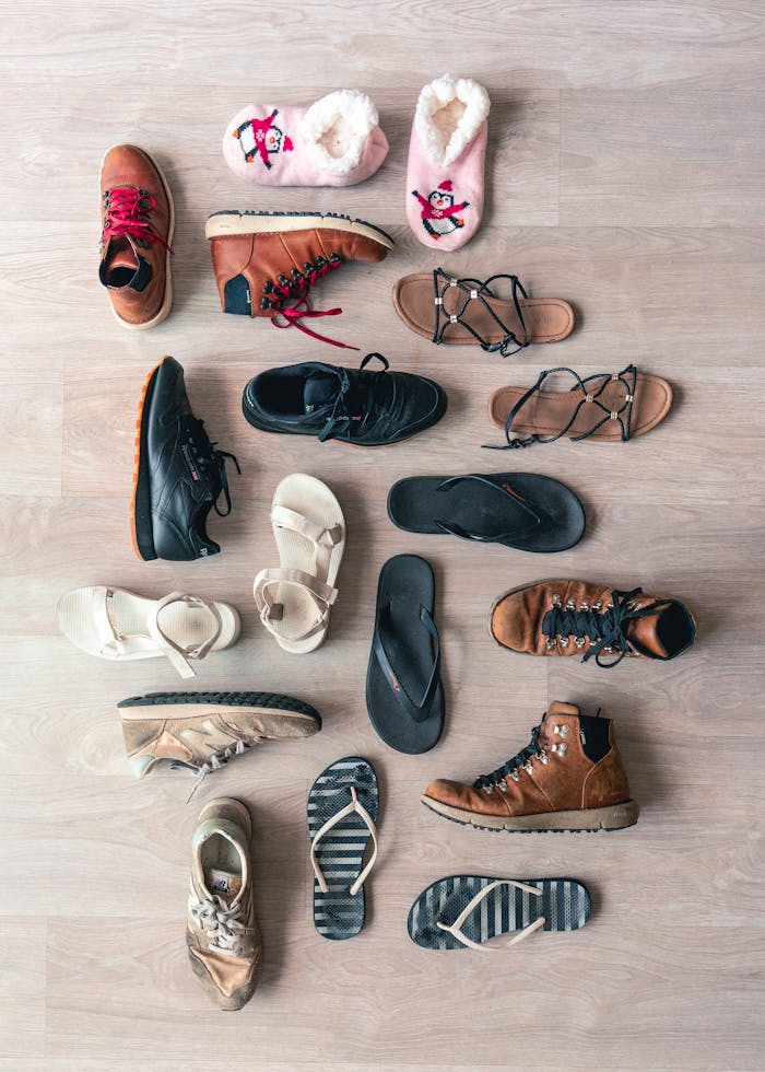 A diverse collection of footwear including boots, sandals, and slippers arranged in a flat lay on a wooden surface.